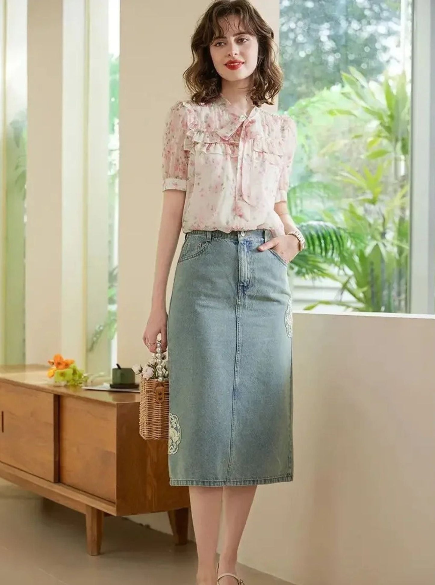 Modell i rosa bluse og skjørt, holder kurvveske ved vinduskarm