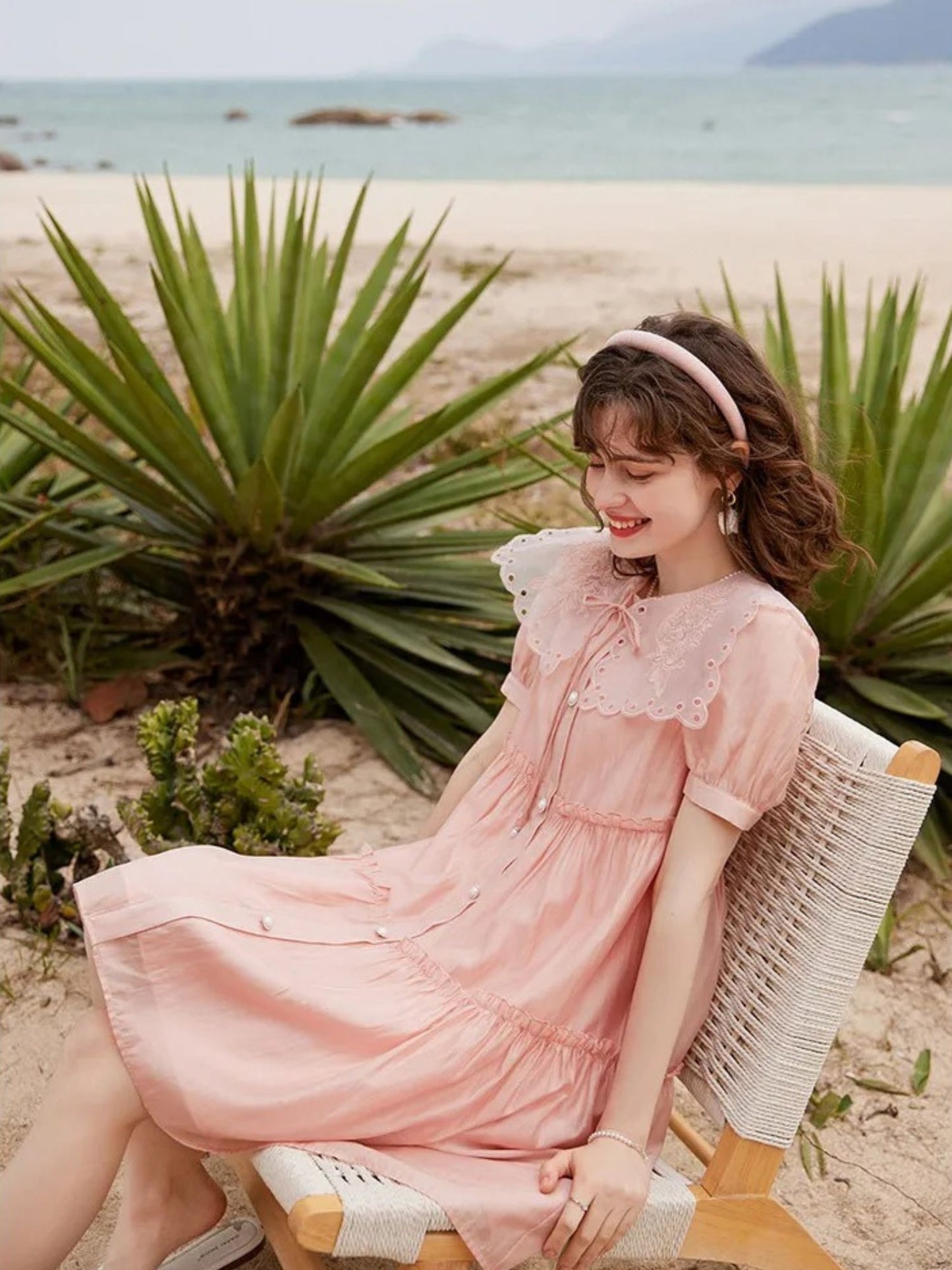 Modell sitter i stol med rosa kjole og hodebånd, ved strandplanter