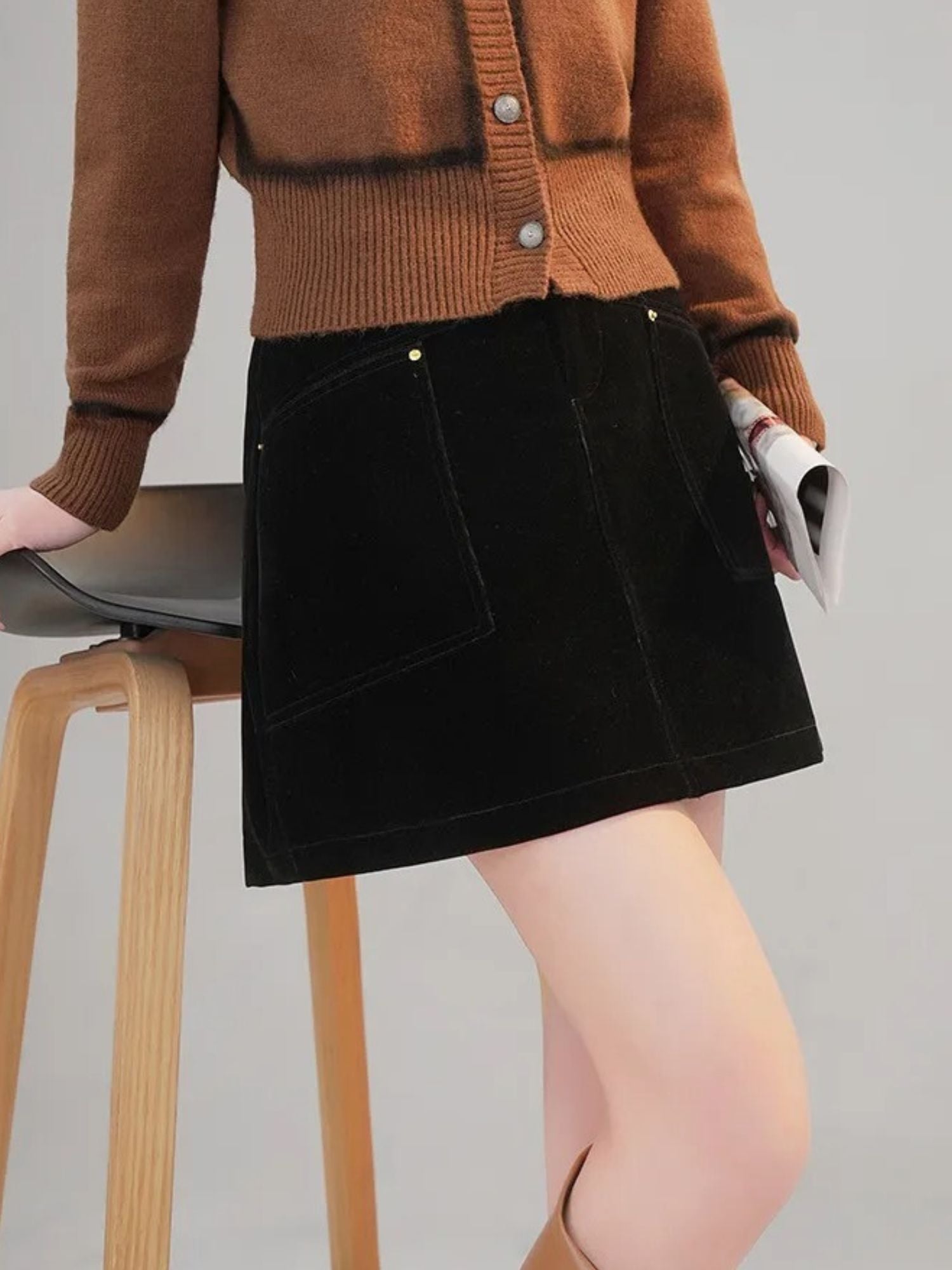 Modell med kort svart kordfløyelsskjørt og brun cardigan, sittende på en stol


